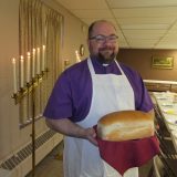 Pastor Greg is wearing a purple clergy shirt and a white apron. He is smiling warmly at the camera and holding a loaf of fresh bread.