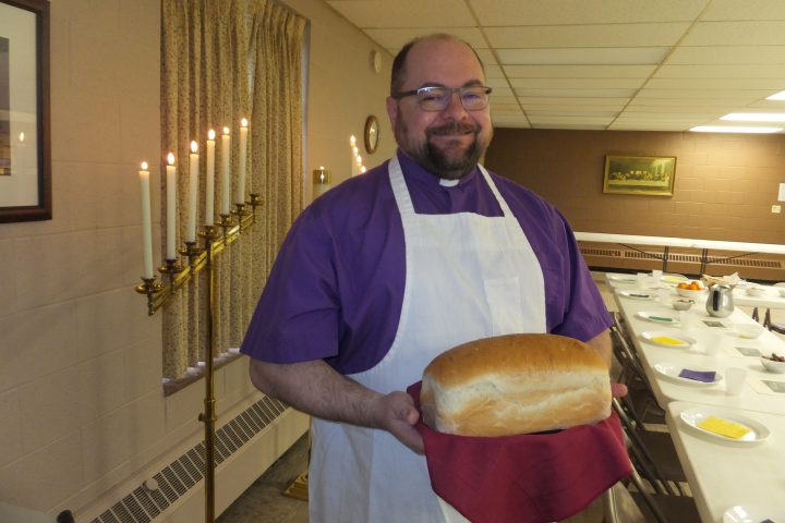 Pastor Greg is wearing a purple clergy shirt and a white apron. He is smiling warmly at the camera and holding a loaf of fresh bread.