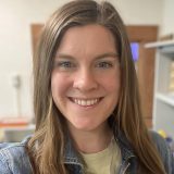 Photo of Tara Downs, a white woman with long brown hair. She is smiling broadly at the camera and wearing a denim jacket over a light green shirt.