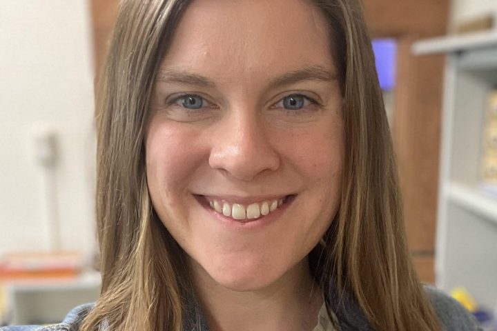 Photo of Tara Downs, a white woman with long brown hair. She is smiling broadly at the camera and wearing a denim jacket over a light green shirt.