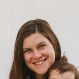 Tara Downs is a white woman with long brown hair. Her head is tilted and she is smiling broadly.