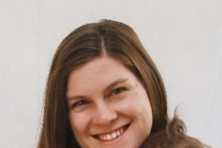 Tara Downs is a white woman with long brown hair. Her head is tilted and she is smiling broadly.